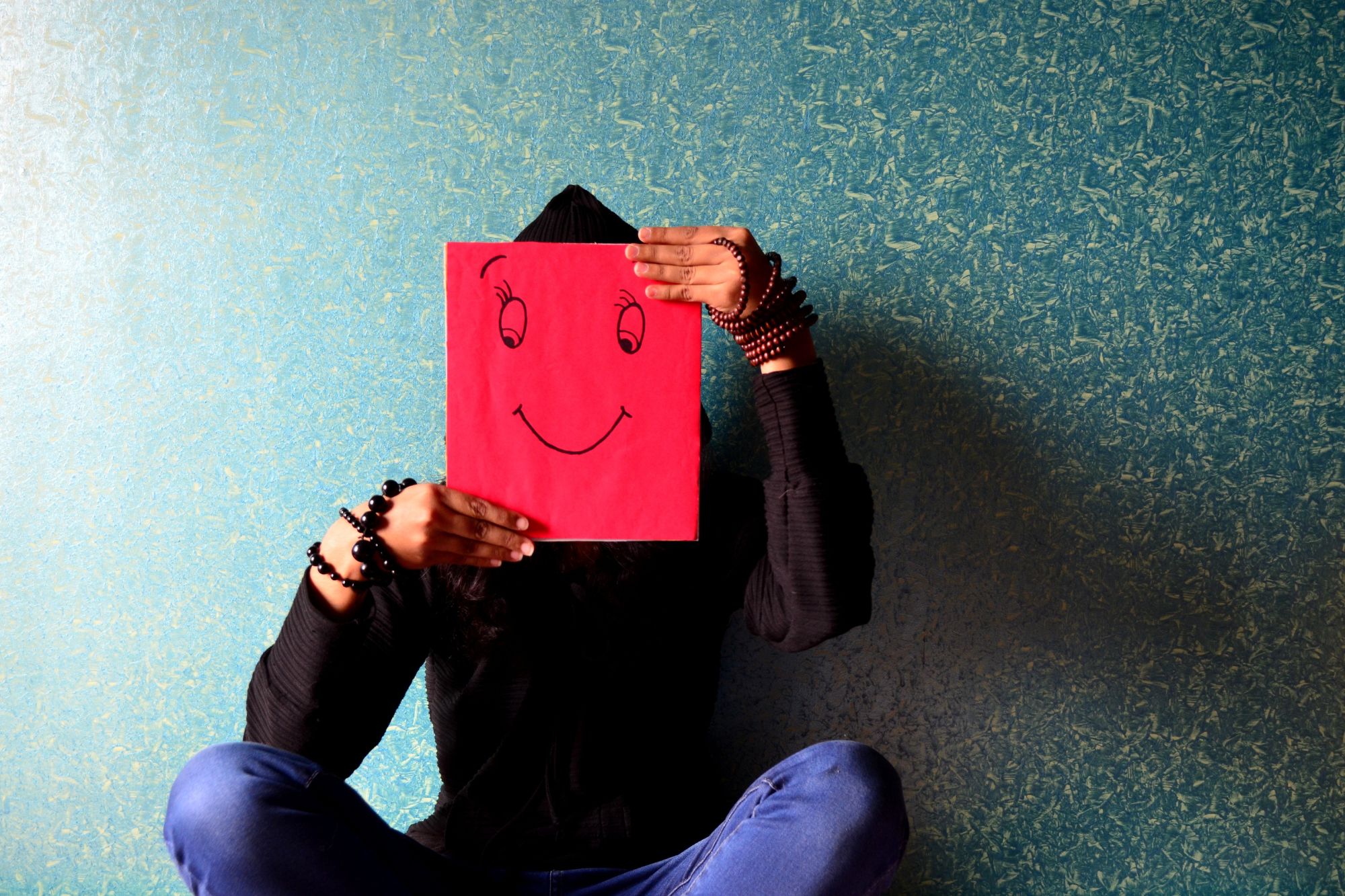 A sad person sitting against a blue background while holding a drawing of a smiley face in front them thinking about how keeping my word to my husband nearly destroyed my life.