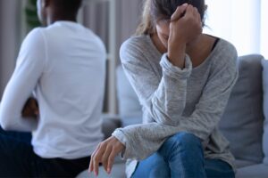 Husband and wife sit turned away from each other as the women cries, wondering why Infidelity Is So Painful To The Betrayed Spouse.